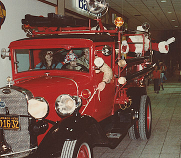Sandy riding in the front seat of an antique fire truck bringing Santa into a mall